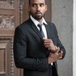 Elegant man in a black suit posing confidently outdoors with a serious expression.